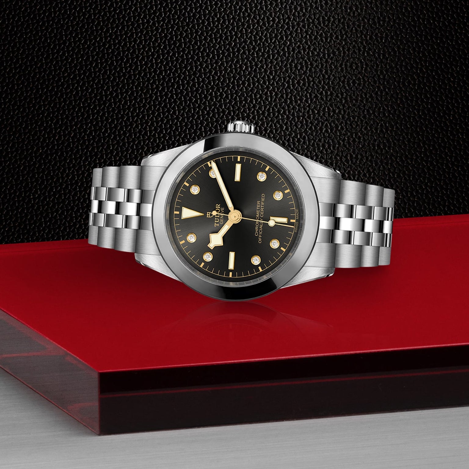 The TUDOR Black Bay One 39, a chic unisex stainless steel wristwatch with a black dial and gold hour markers, rests on a glossy red and black surface, reflecting its refined design against a textured black background.