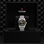 The TUDOR Black Bay One 39 wristwatch with a black dial and silver bracelet is upright in an open black box featuring the TUDOR logo, highlighting its unisex chic style and refined elegance.