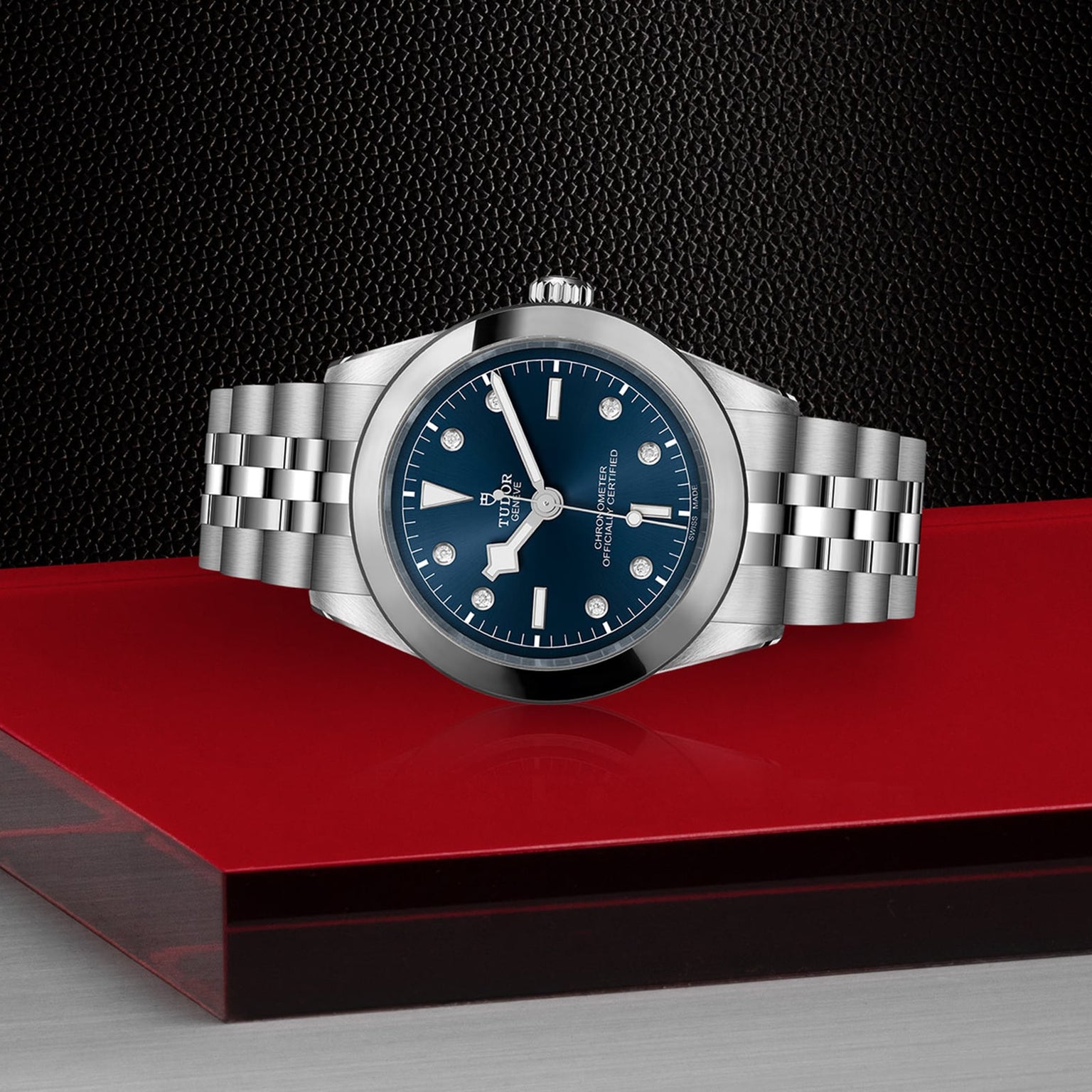 The TUDOR Black Bay One 39 is a unisex stainless steel wristwatch with a blue dial and silver link bracelet, displayed on red against a textured black backdrop. Inspired by Black Bay models, it offers timeless elegance and versatility.