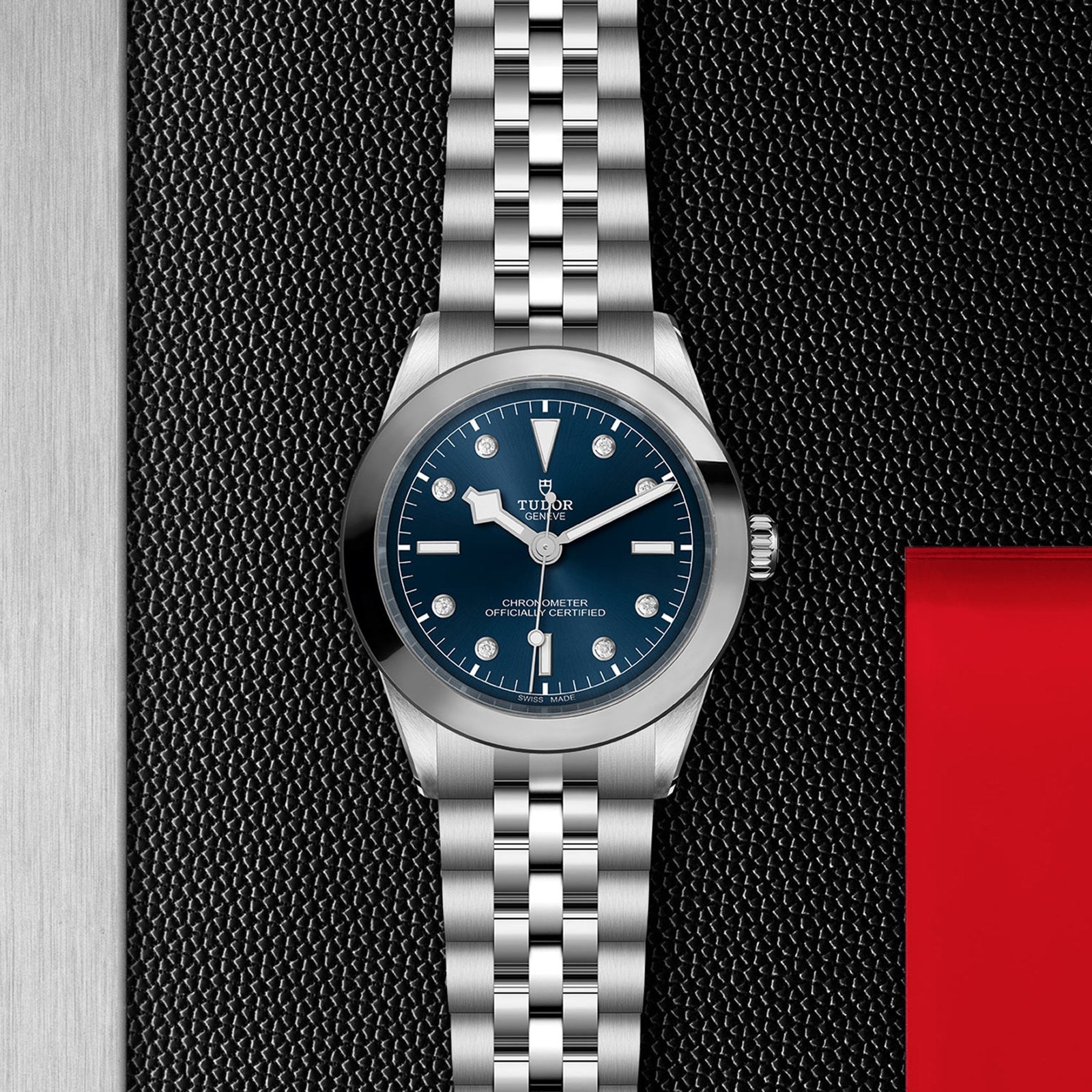 The TUDOR Black Bay One 39 is a unisex stainless steel watch with a blue dial and silver bracelet, showcased on a textured black surface with silver and red accents—reflecting the iconic Black Bay design.