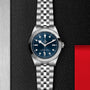 The TUDOR Black Bay One 39 is a unisex stainless steel watch with a blue dial and silver bracelet, showcased on a textured black surface with silver and red accents—reflecting the iconic Black Bay design.