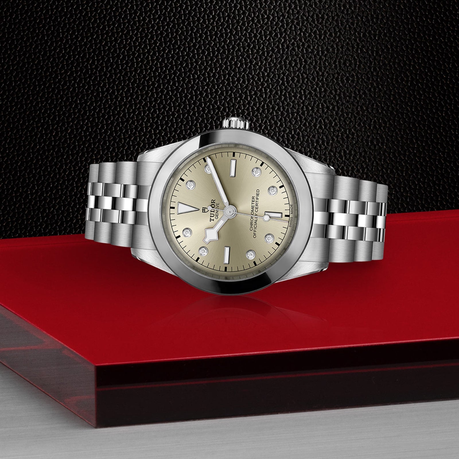 The TUDOR Black Bay One 39 unisex watch with a gold dial, diamond hour markers, and a 316L steel bracelet is displayed on a red and black geometric surface.