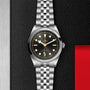 The TUDOR Black Bay One 41 wristwatch with a black dial and jubilee bracelet is displayed on a textured black surface.