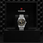 The TUDOR Black Bay One 41, a silver wristwatch with a black dial and gold accents, is presented in an open black TUDOR box with logo branding, exuding classic unisex style inspired by Black Bay models.