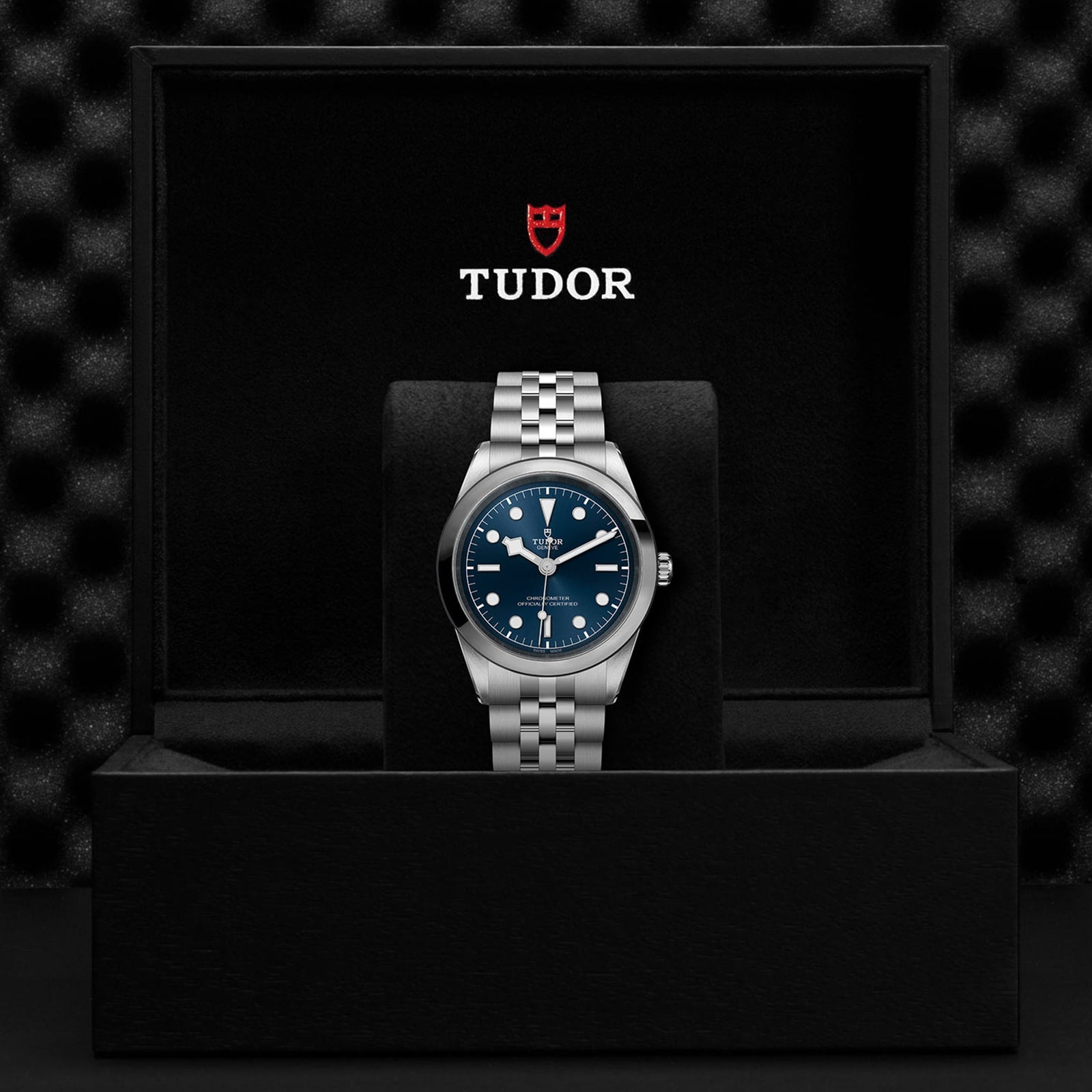 A TUDOR Black Bay One 41 wristwatch with a blue dial and silver bracelet is showcased upright in a black presentation box with the TUDOR logo, highlighting its unisex chic style.
