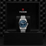 A TUDOR Black Bay One 41 wristwatch with a blue dial and silver bracelet is showcased upright in a black presentation box with the TUDOR logo, highlighting its unisex chic style.