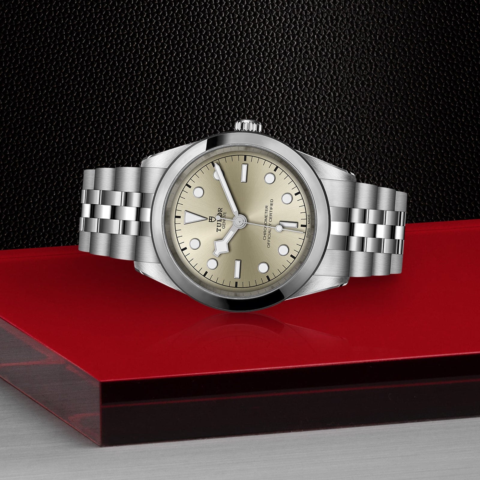 A TUDOR Black Bay One 41 wristwatch with a light gold dial and metal bracelet sits on red glass, radiating unisex style against a textured black backdrop.