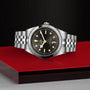 The TUDOR Black Bay One 41, a unisex chic wristwatch with a black dial and gold markers, is crafted from premium 316L stainless steel and displayed on a red and gray platform against a textured black background.