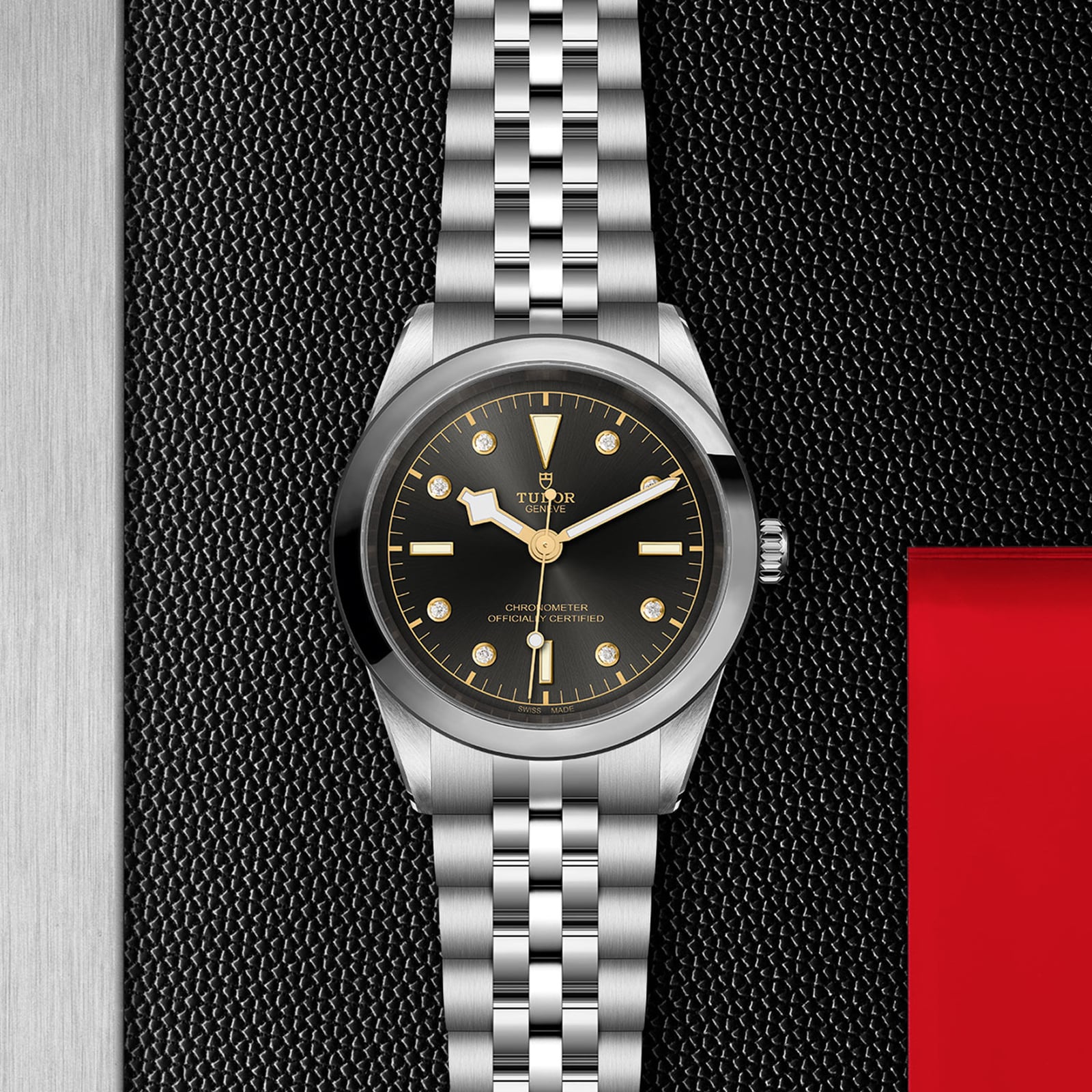 The TUDOR Black Bay One 41, a unisex wristwatch made from 316L steel with a black dial and silver bracelet, is displayed on a textured black and red background.
