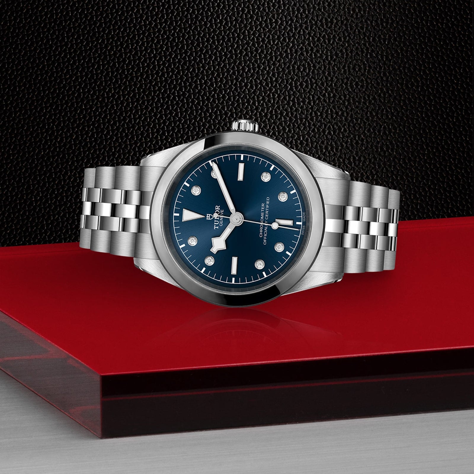The TUDOR Black Bay One 41, a unisex chic stainless steel wristwatch with a blue dial, is showcased on a red and black platform set against a textured black background, reflecting the refined Black Bay design.