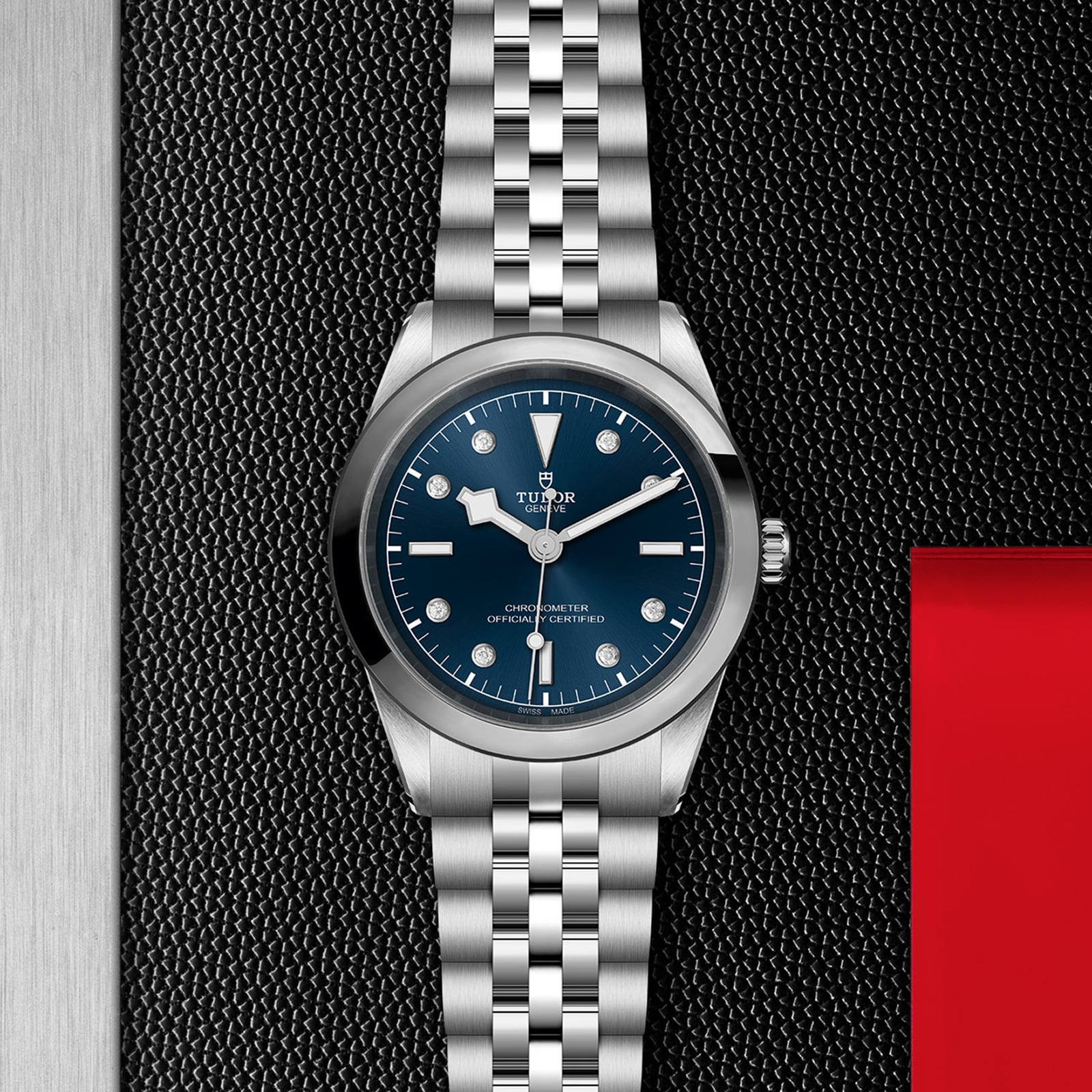 The TUDOR Black Bay One 41, a silver watch with a blue dial and metal bracelet featuring the TUDOR T fit adjustment system, is displayed on a textured black surface with a red accent, embodying unisex chic.