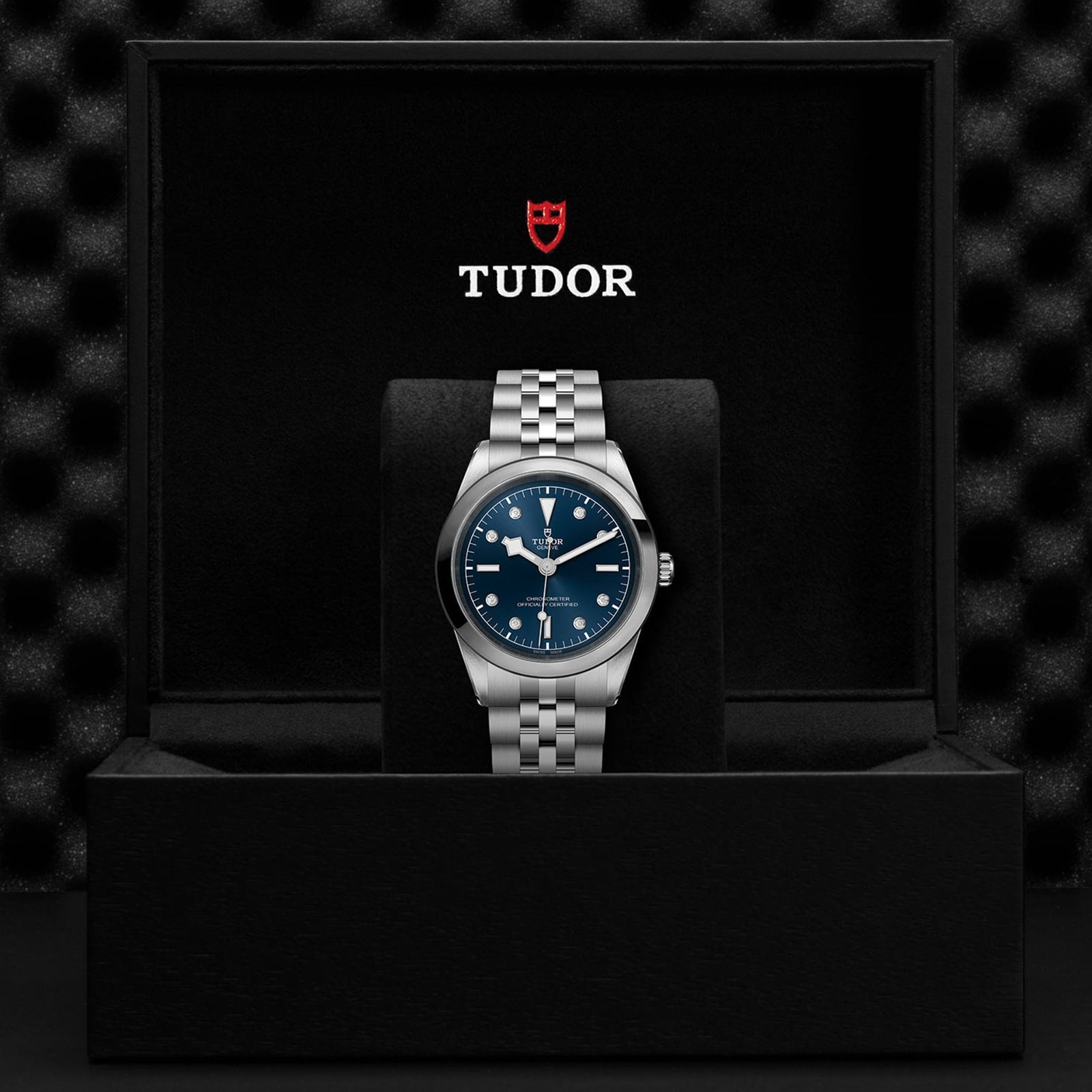 The TUDOR Black Bay One 41, featuring a blue dial and silver bracelet, is showcased upright in a black branded box with the TUDOR logo above it and highlighted by the innovative unisex T fit adjustment system.