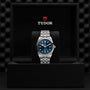 The TUDOR Black Bay One 41, featuring a blue dial and silver bracelet, is showcased upright in a black branded box with the TUDOR logo above it and highlighted by the innovative unisex T fit adjustment system.