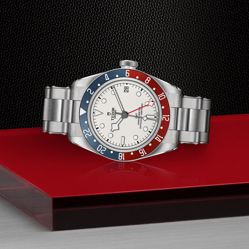 The TUDOR Black Bay GMT, featuring a silver case, red and blue bezel, opaline dial, and stainless steel band, is displayed on a red and black surface with a textured black background—a striking tribute to its iconic design.
