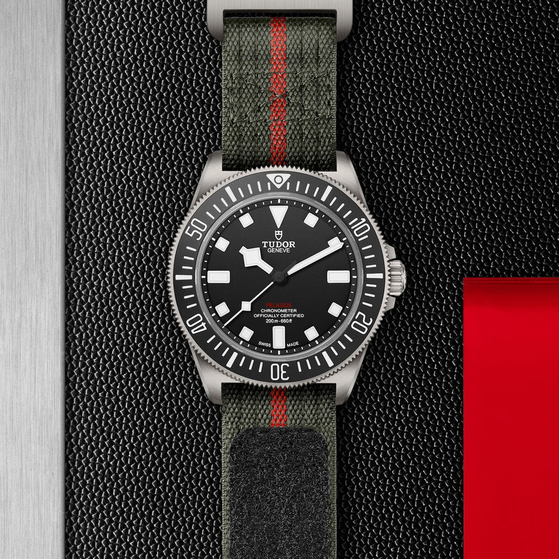The TUDOR Pelagos FXD wristwatch features a black dial, white markers, and a green fabric strap with red stripe, set against a textured black and red backdrop—showcasing the heritage of TUDOR diving watches.