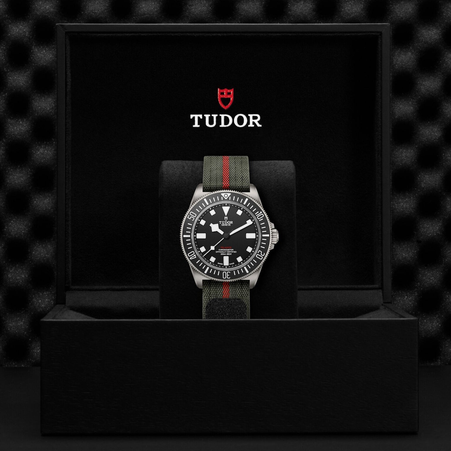 The TUDOR Pelagos FXD, inspired by Military Submariner heritage, features a black dial and striped fabric strap, displayed upright in a black presentation box with the TUDOR logo on the inner lid.