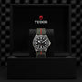 The TUDOR Pelagos FXD, inspired by Military Submariner heritage, features a black dial and striped fabric strap, displayed upright in a black presentation box with the TUDOR logo on the inner lid.