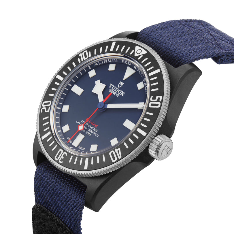 The TUDOR Pelagos FXD features a blue dial with white markers, black case, and fabric strap. Designed for underwater navigation, this watch offers a waterproof build and is shown angled for detail.