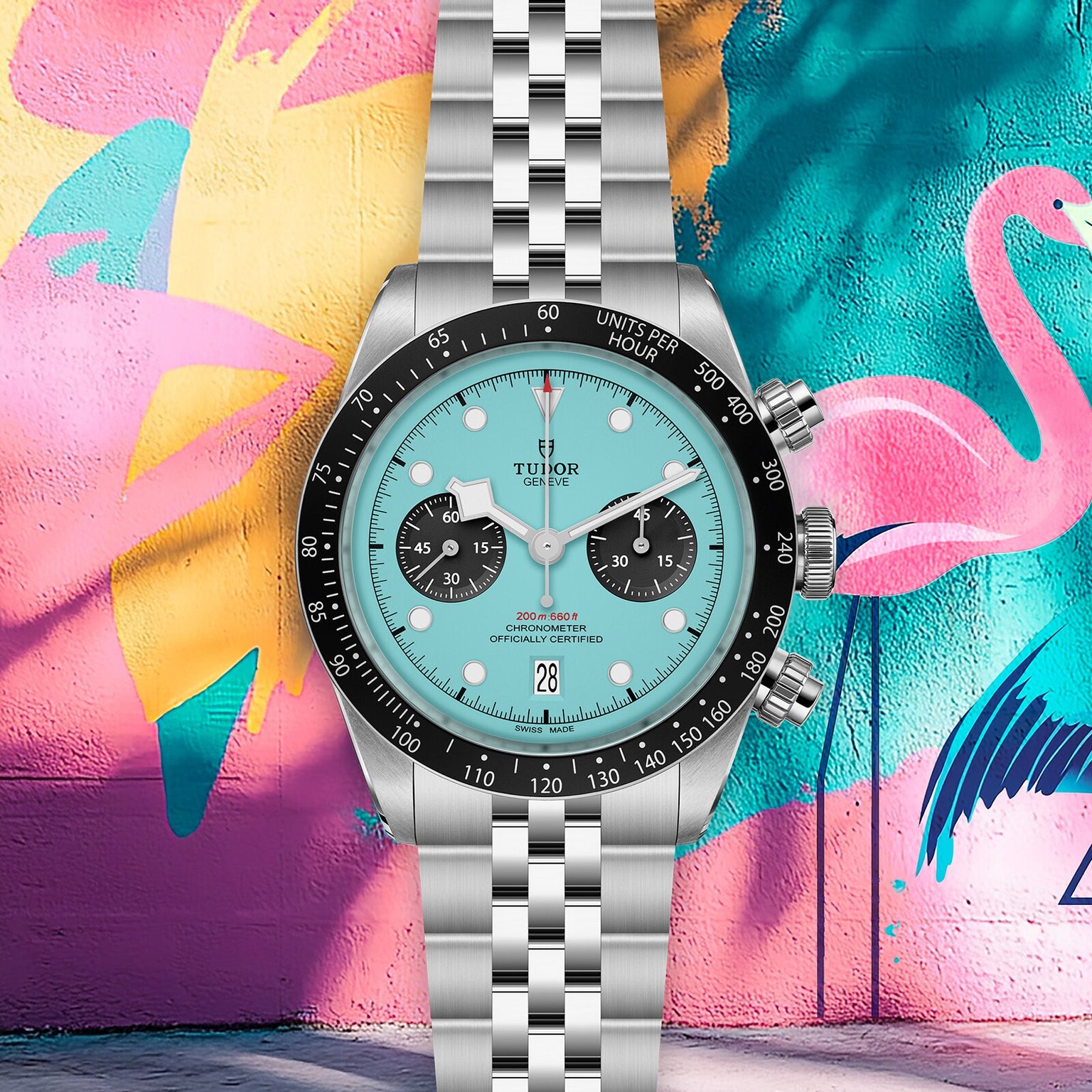 A TUDOR Black Bay Chrono wristwatch with a turquoise dial and black subdials is showcased before a vibrant wall mural with a flamingo and abstract shapes.