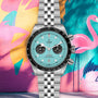 A TUDOR Black Bay Chrono wristwatch with a turquoise dial and black subdials is showcased before a vibrant wall mural with a flamingo and abstract shapes.