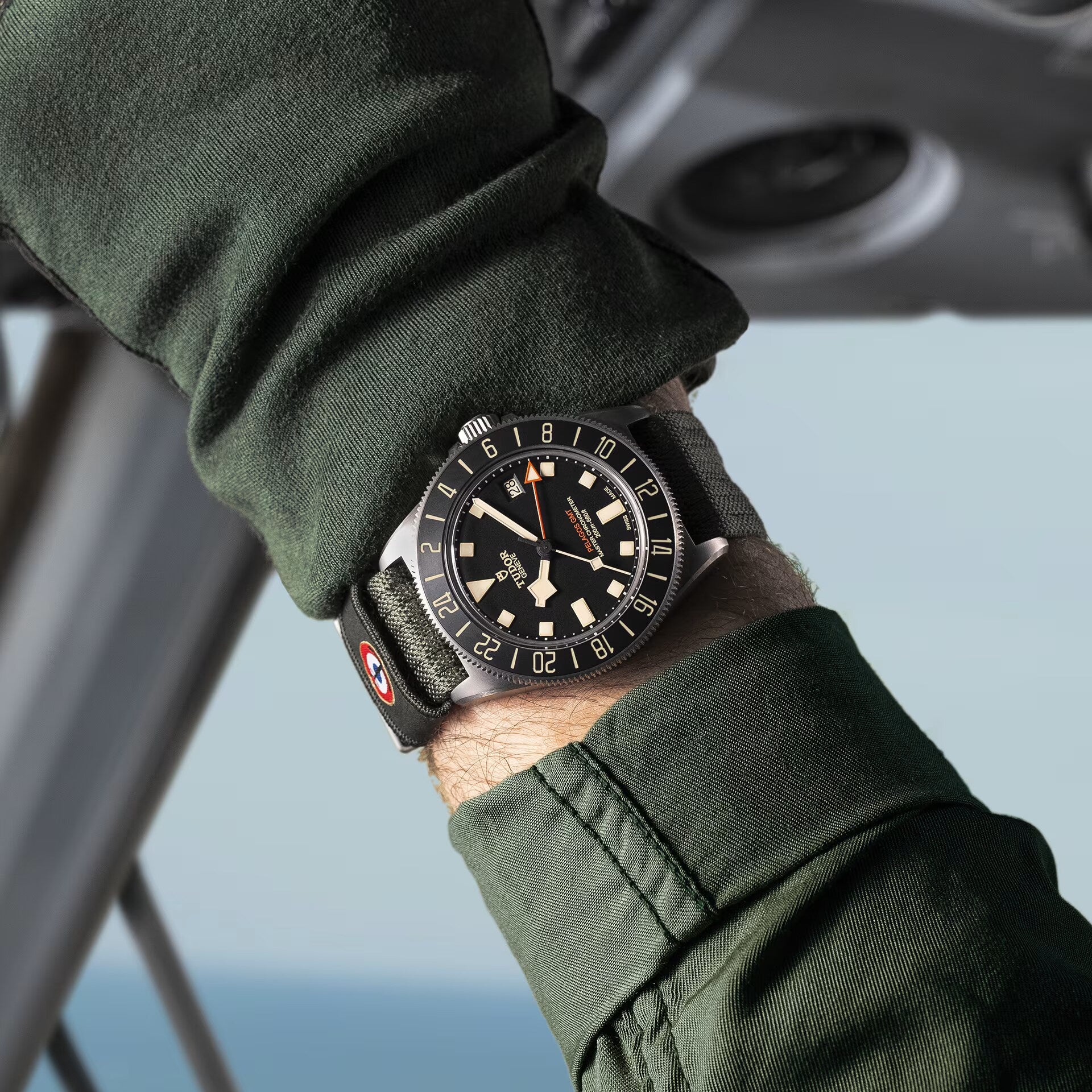 A person in a dark green jacket showcases the TUDOR Pelagos FXD GMT, a black wristwatch with a rotating bezel and fabric strap.