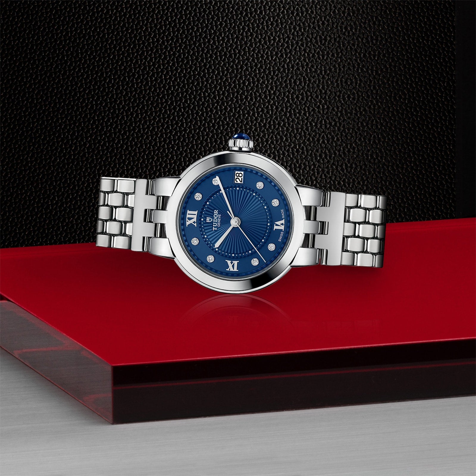The TUDOR Clair de Rose silver wristwatch features a blue face with Roman numerals, displayed on a red and black geometric surface.