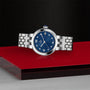 The TUDOR Clair de Rose silver wristwatch features a blue face with Roman numerals, displayed on a red and black geometric surface.
