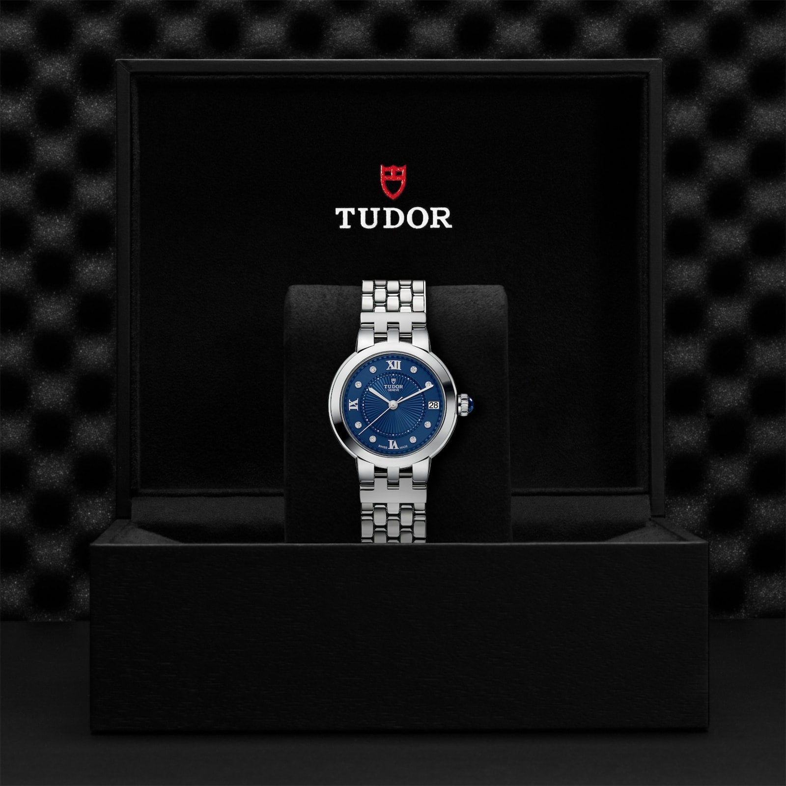 A silver TUDOR Clair de Rose wristwatch with a blue face, presented in a black TUDOR-branded display box.