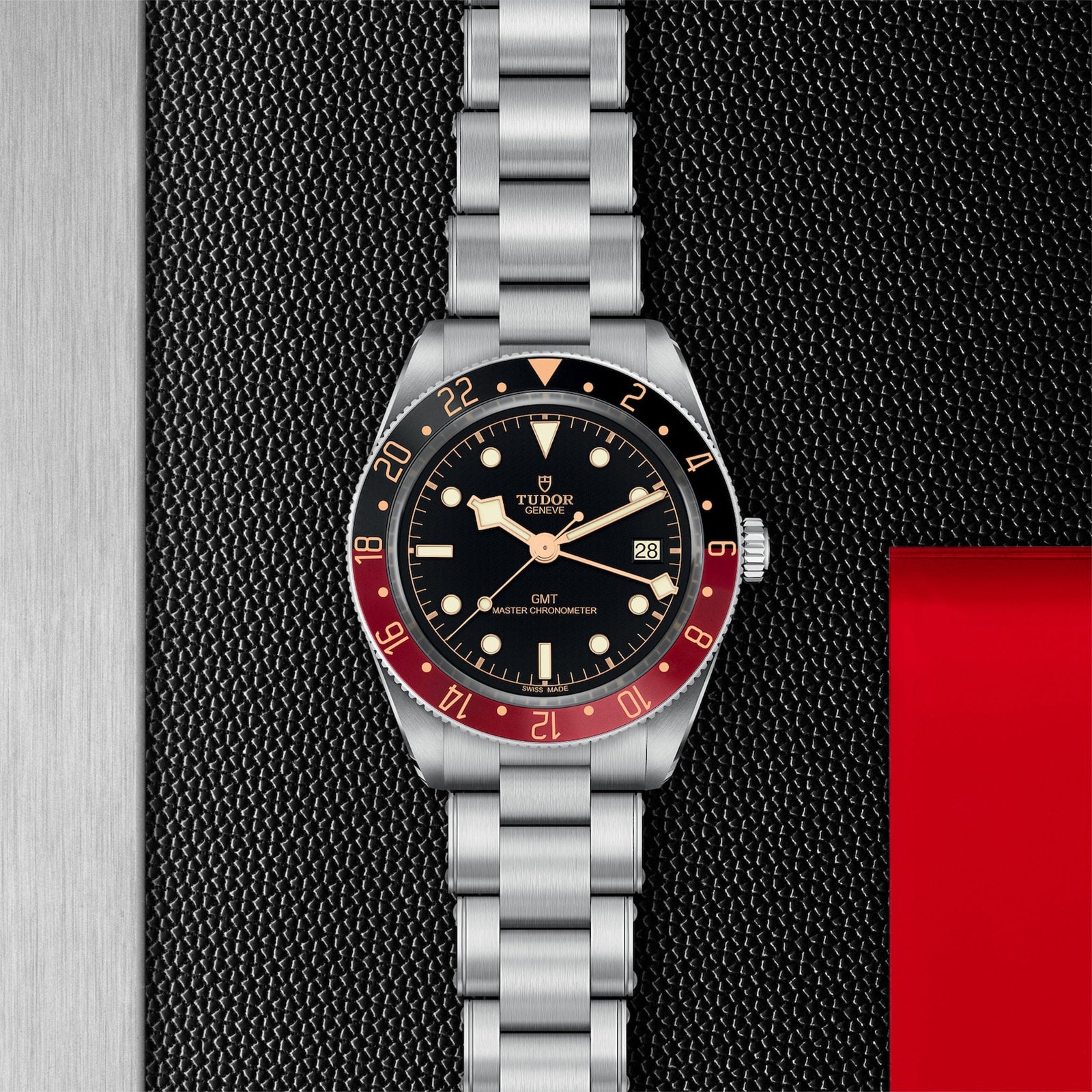 A TUDOR Black Bay 58 GMT dive watch with a black dial, red and black bezel, and metal bracelet sits on a textured black surface featuring a metallic edge and red section.