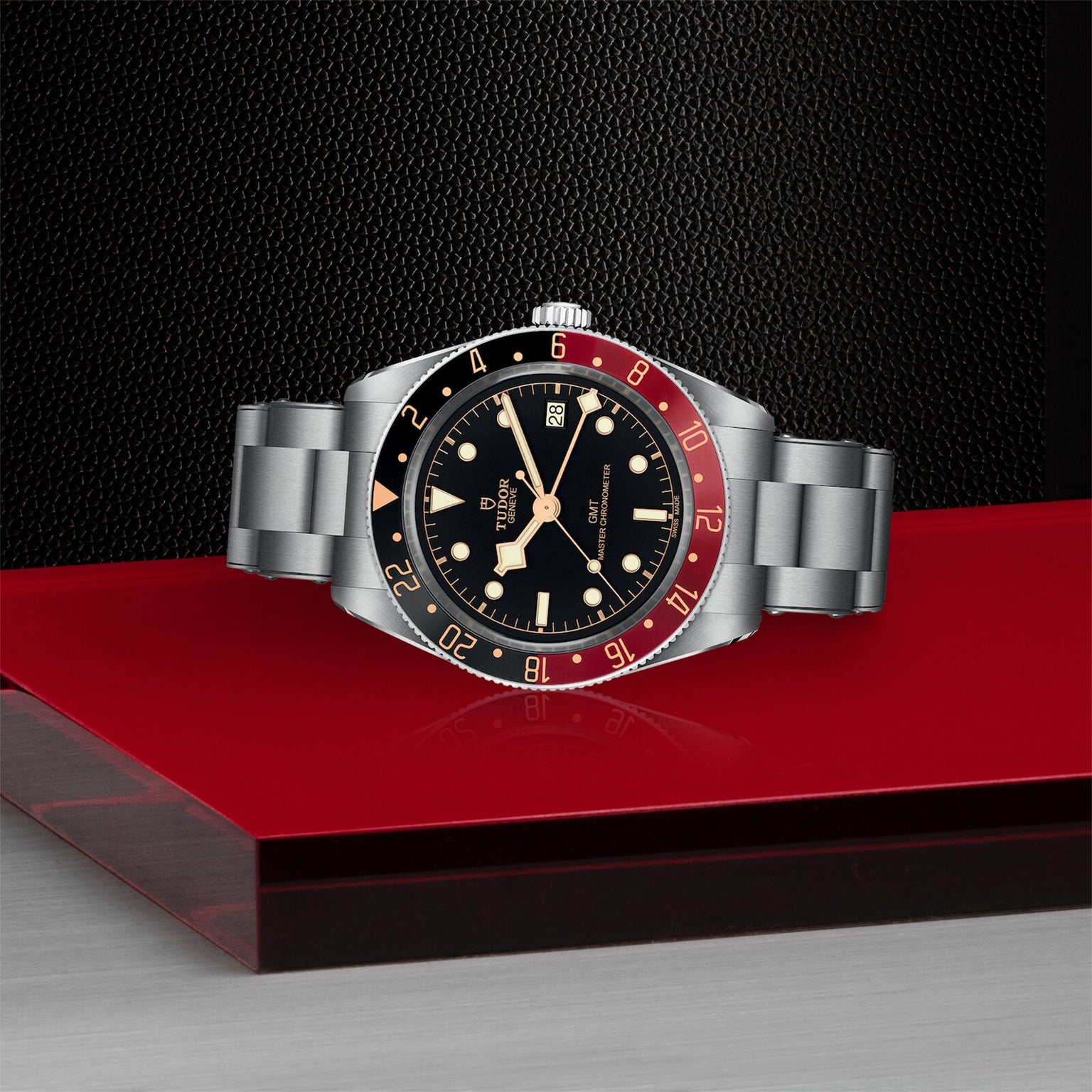 A TUDOR Black Bay 58 GMT watch with a black dial, red and black bezel, and metal bracelet rests on a red and silver surface against a textured black background.