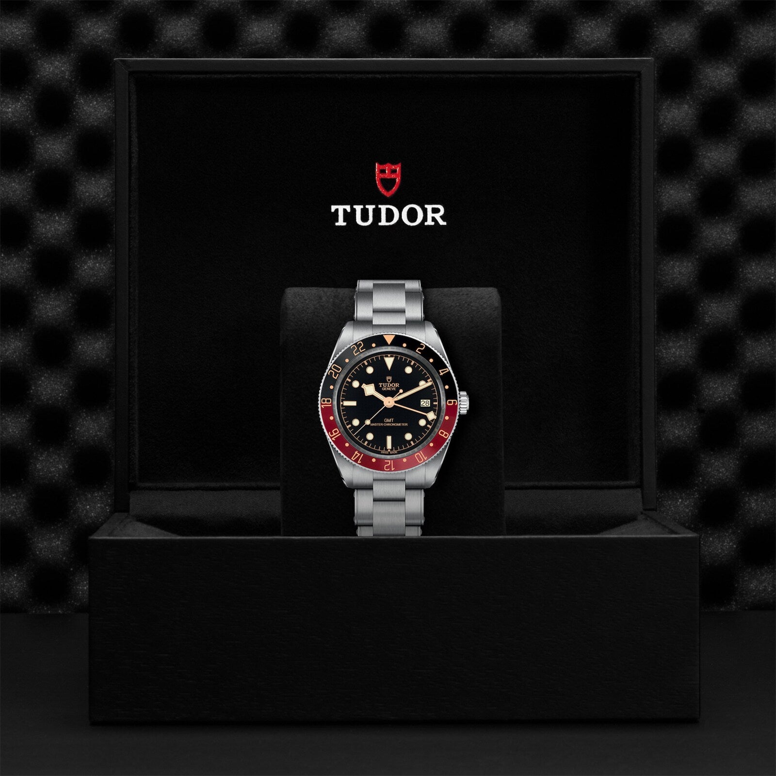 A TUDOR Black Bay 58 GMT wristwatch with a black and red bezel is shown in an open black box featuring the TUDOR logo on the inside lid.
