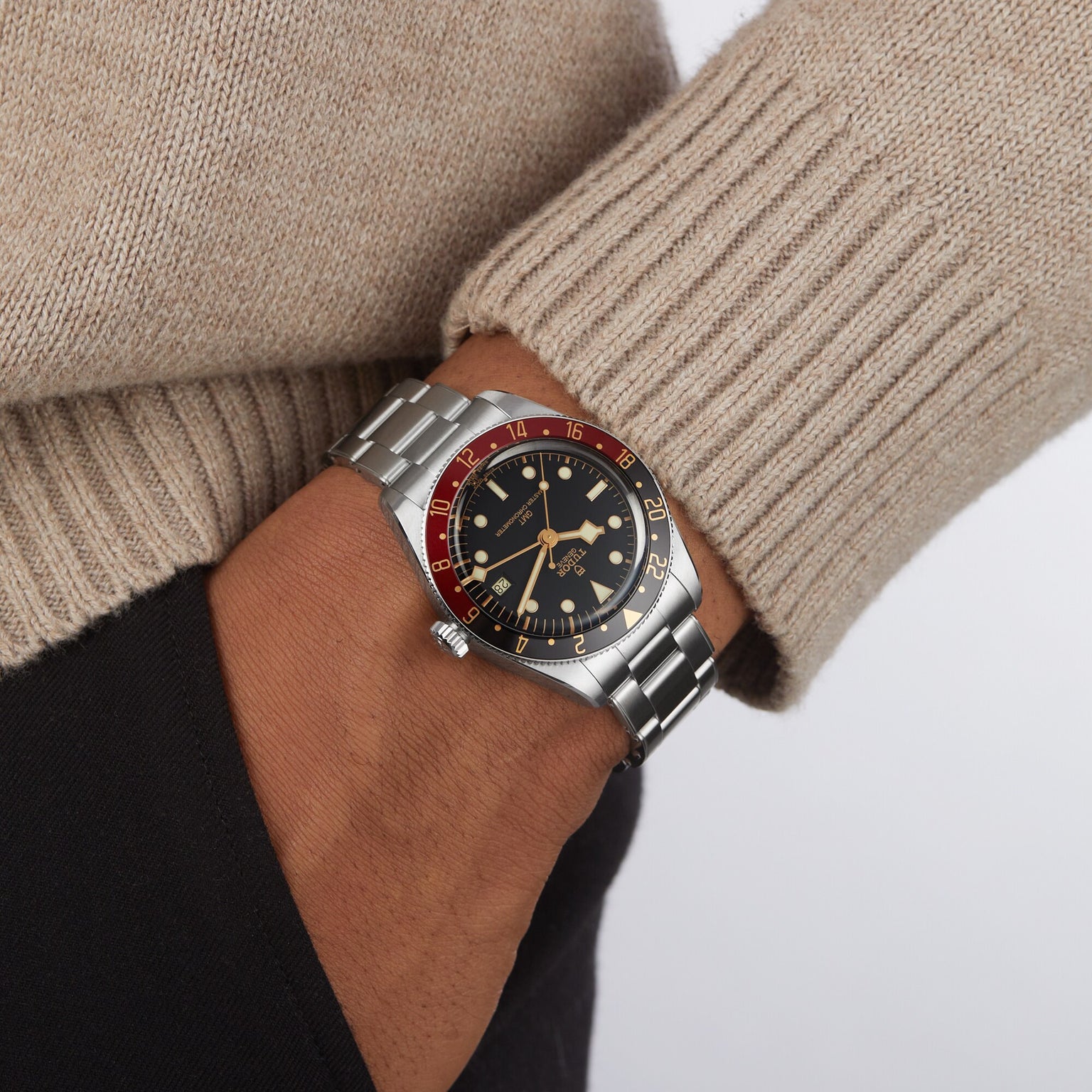 A person in a beige sweater and black pants wears a TUDOR Black Bay 58 GMT dive watch with a black and red bezel and black dial on their left wrist.