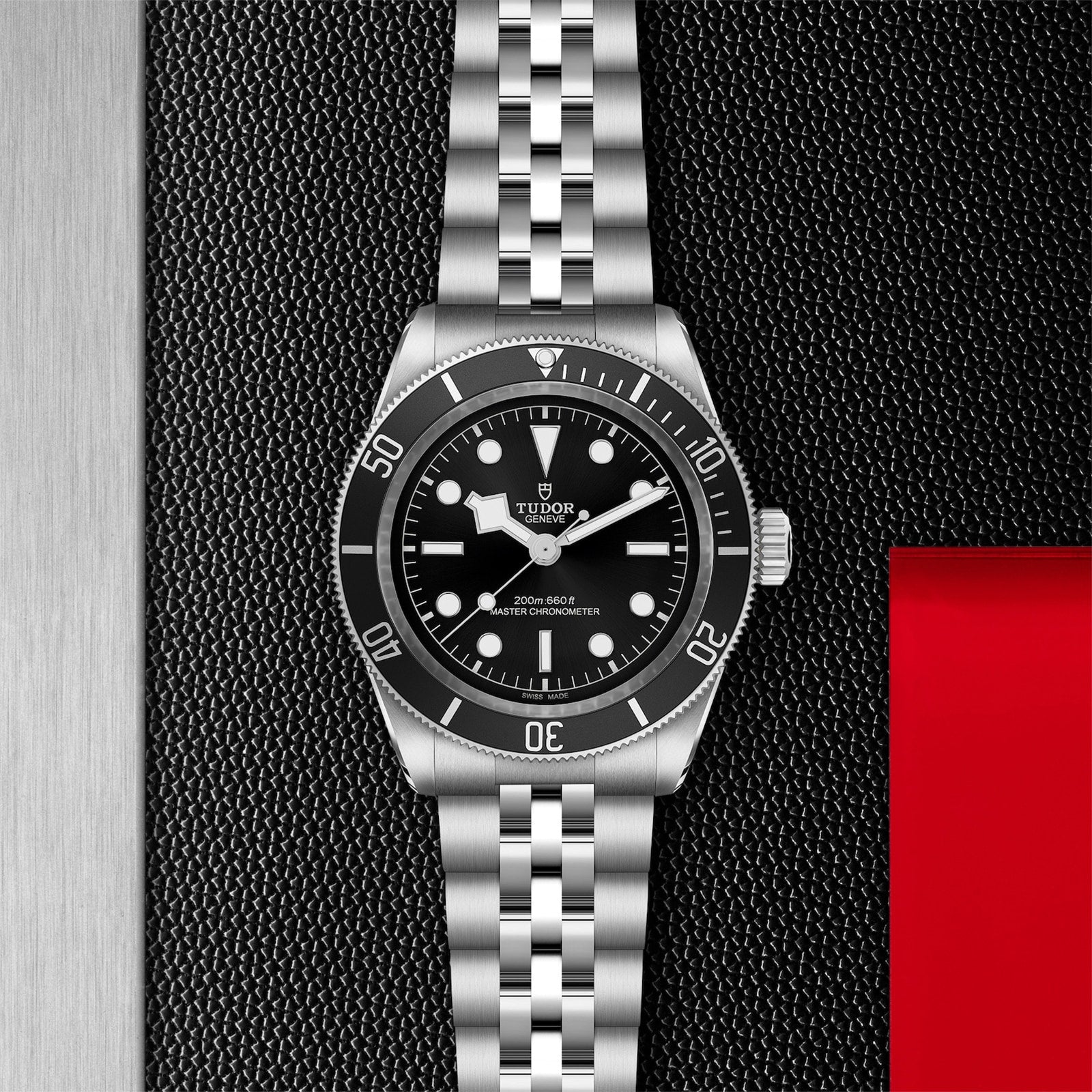 A TUDOR Black Bay wristwatch with a black dial and metal bracelet is displayed on a textured black surface beside red and silver panels, showcasing the Black Bays heritage as an iconic diving watch.