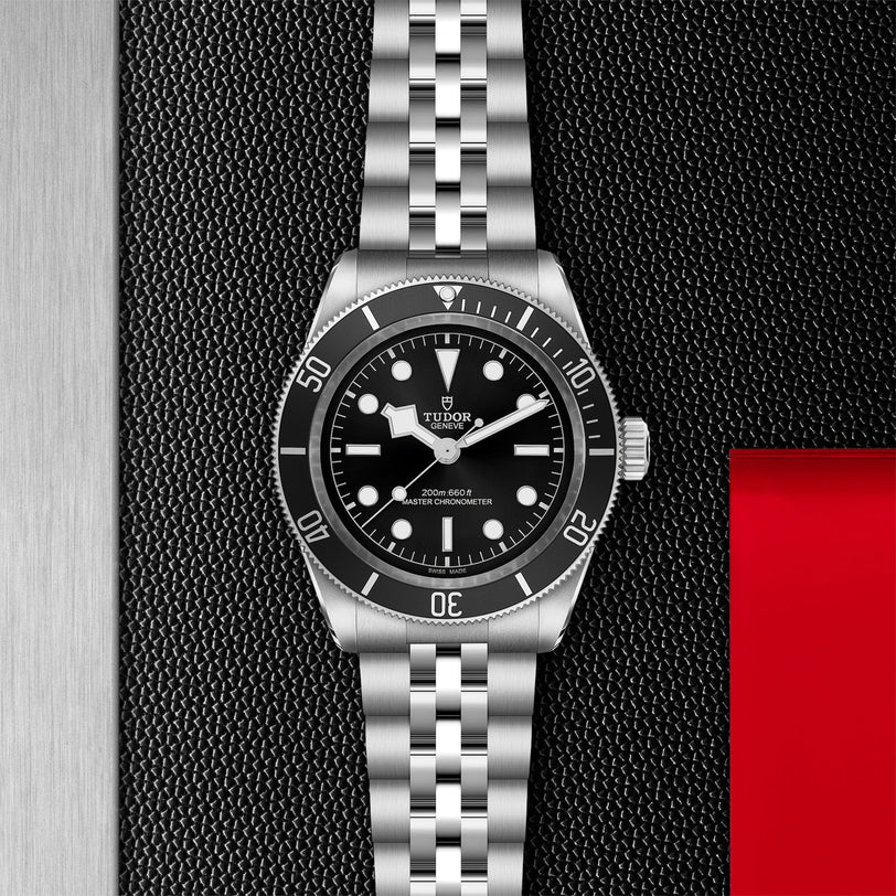A TUDOR Black Bay wristwatch with a black dial and metal bracelet is displayed on a textured black surface beside red and silver panels, showcasing the Black Bays heritage as an iconic diving watch.