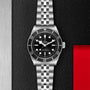 A TUDOR Black Bay wristwatch with a black dial and metal bracelet is displayed on a textured black surface beside red and silver panels, showcasing the Black Bays heritage as an iconic diving watch.