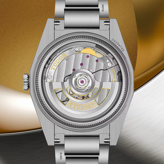 Stainless steel TUDOR Monarch watch with exposed mechanical movement, gold accents, and brushed metal bracelet against gold a