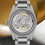 Stainless steel TUDOR Monarch watch with exposed mechanical movement, gold accents, and brushed metal bracelet against gold a