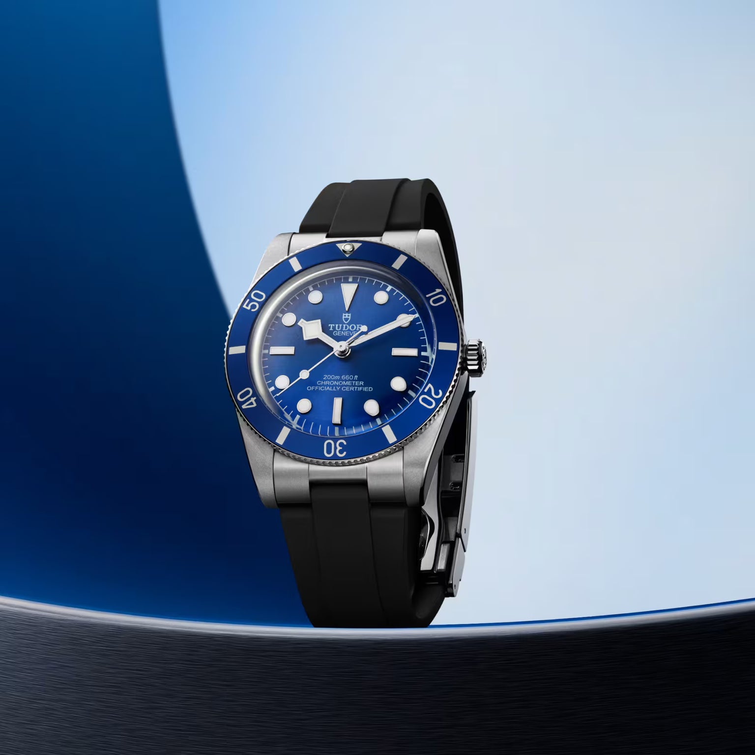 Tudor Black Bay 54 watch with blue dial, silver case, and black rubber strap on blue background.