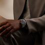 Black watch with dark dial and metal bracelet worn on wrist over brown sleeve.