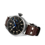 The IWC Schaffhausen Big Pilots Watch Heritage is a silver wristwatch featuring a black dial, blue second hand, and a brown leather strap with metal rivets.