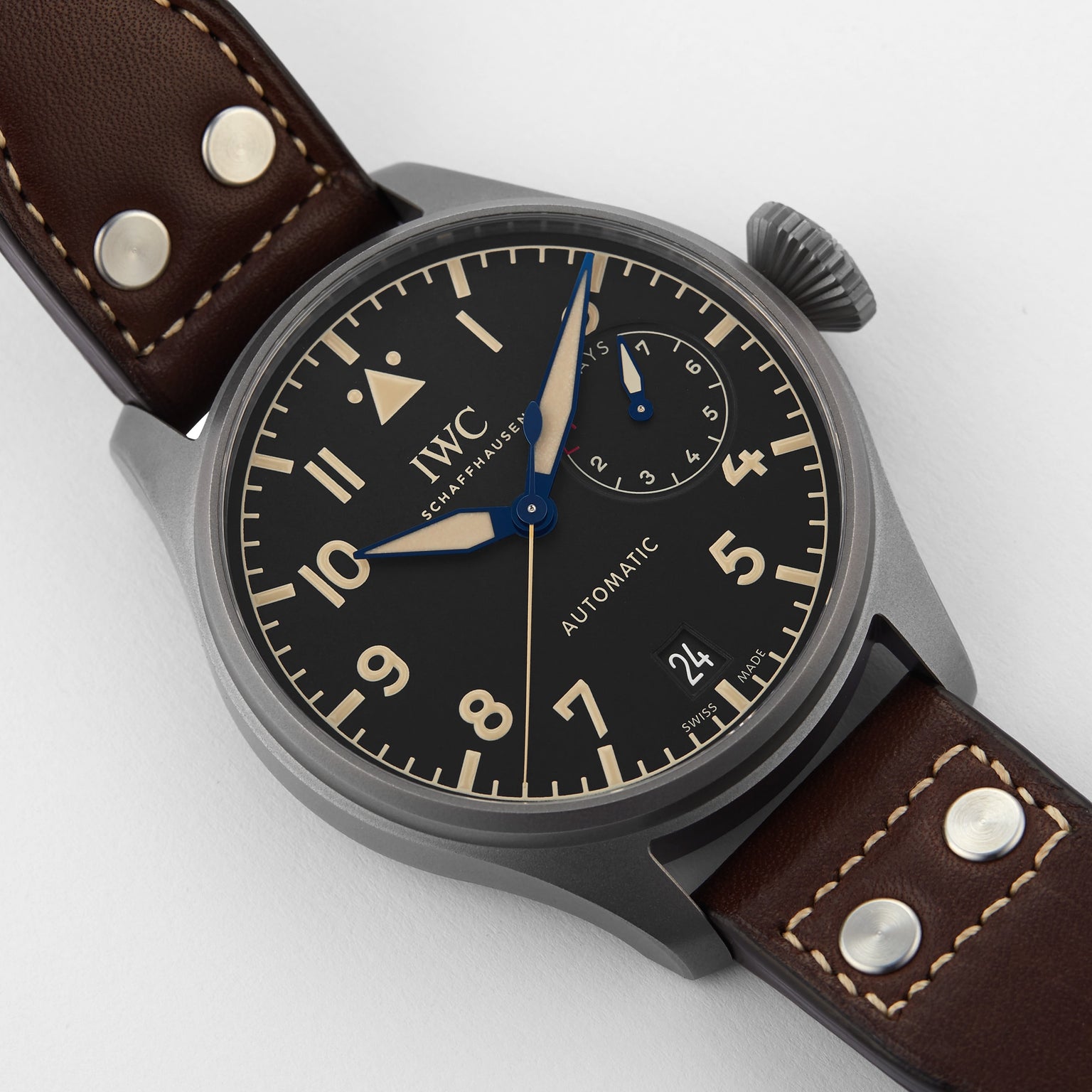A close-up of the IWC Schaffhausen Big Pilots Watch Heritage shows its automatic movement, black dial, brown leather strap, blue power reserve hand, and a date display at 6 oclock.