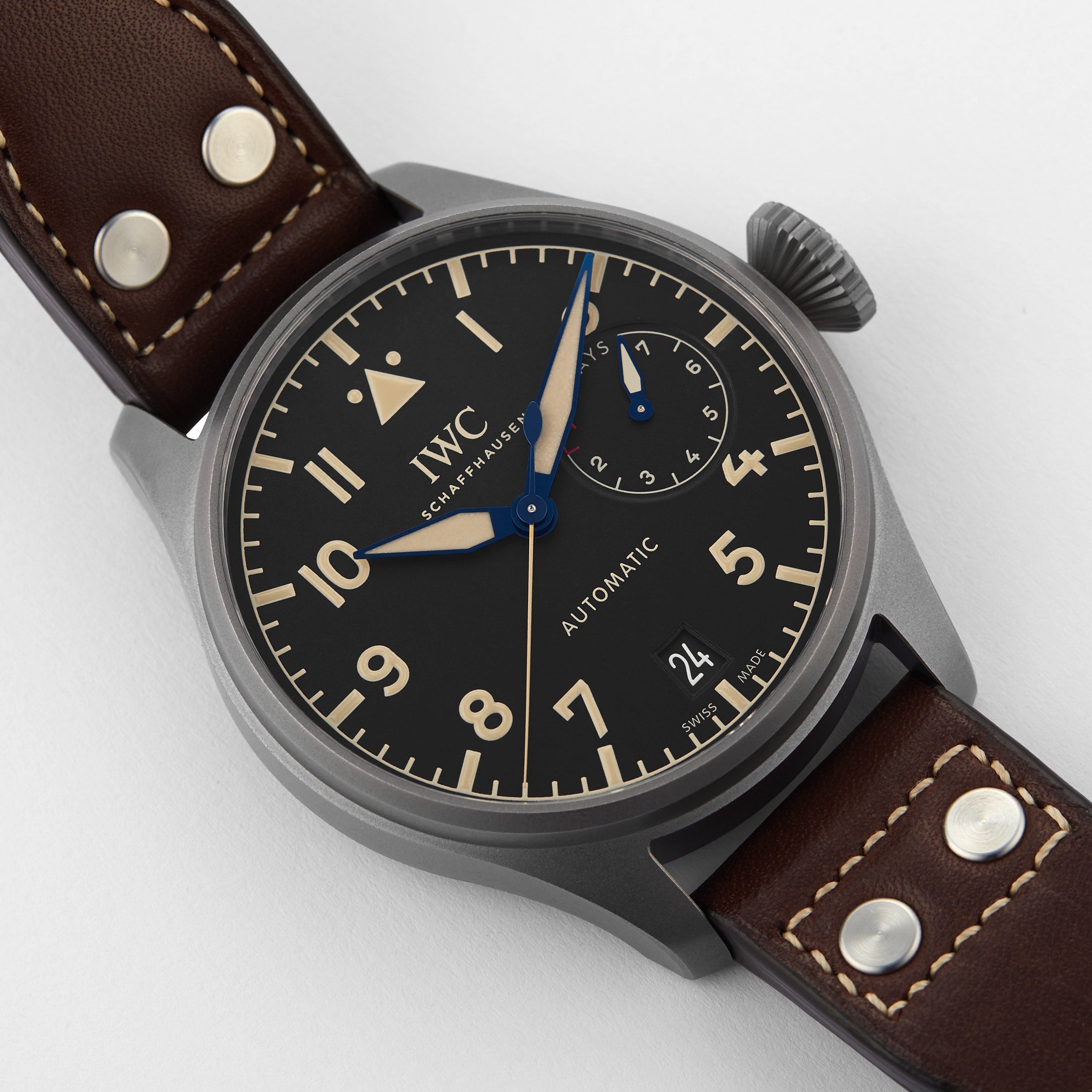 A close-up of the IWC Schaffhausen Big Pilots Watch Heritage shows its automatic movement, black dial, brown leather strap, blue power reserve hand, and a date display at 6 oclock.