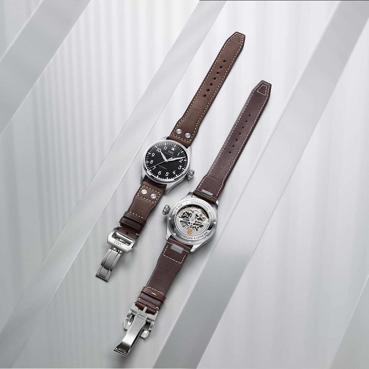 Two IWC Schaffhausen Big Pilot’s Watch 43 timepieces with brown leather straps are displayed—one features a bold black dial, the other showcases its mechanical movement and stainless steel case—on a light-striped background.