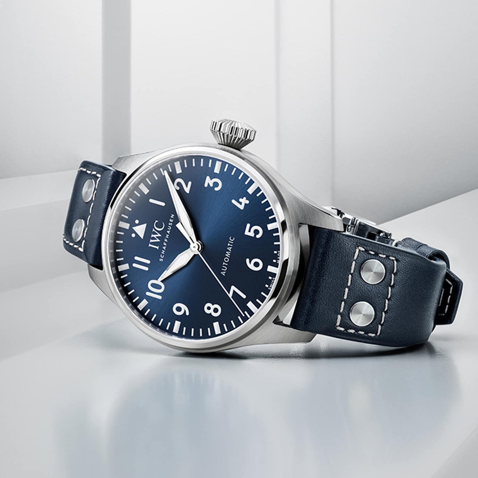 An IWC Schaffhausen Big Pilots Watch 43 with a blue dial, white numerals, and a blue leather strap with metal rivets is displayed on a light gray surface.