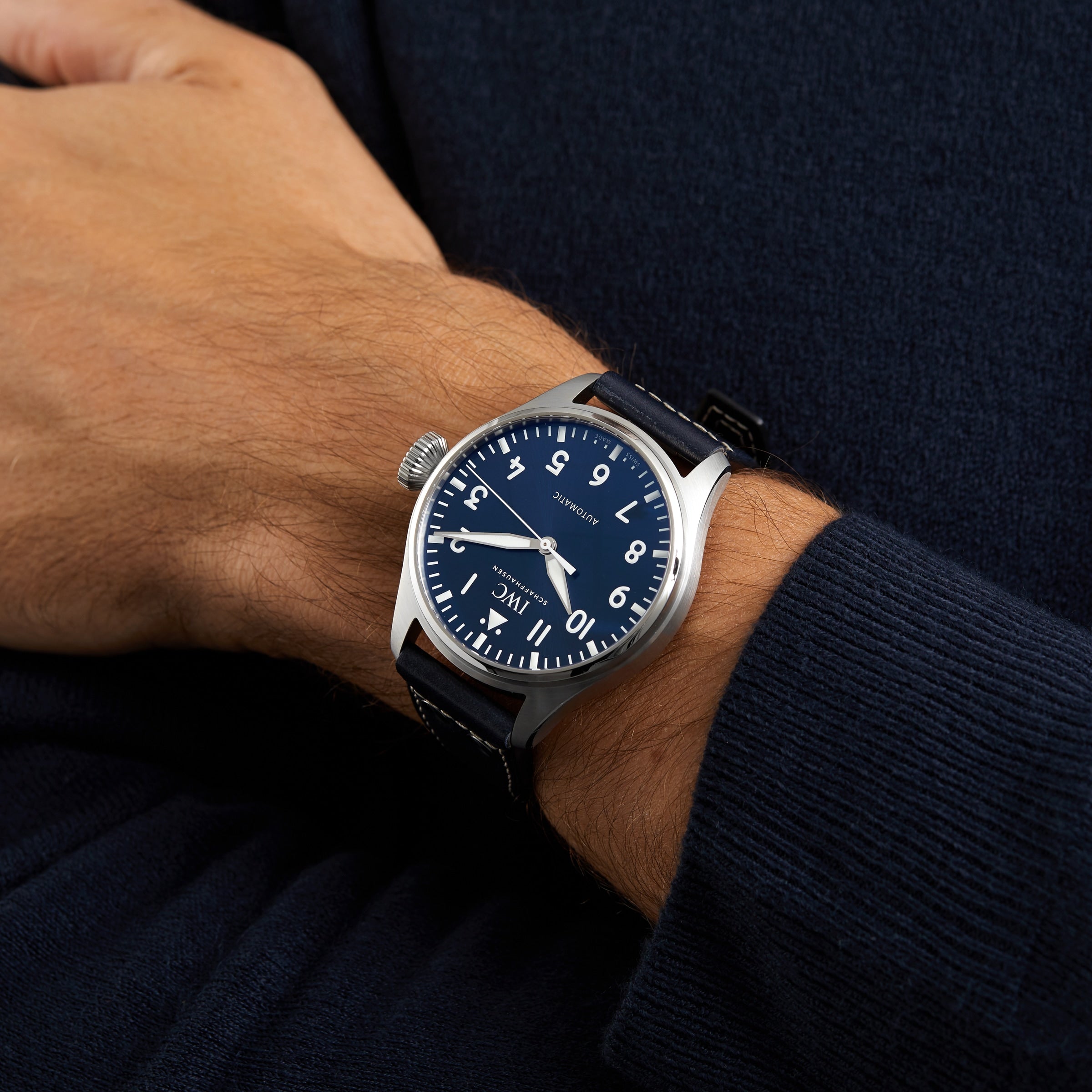 A wrist wearing the IWC Schaffhausen Big Pilots Watch 43 with a blue dial, white numerals, and a black leather strap, shown against a dark blue sweater.