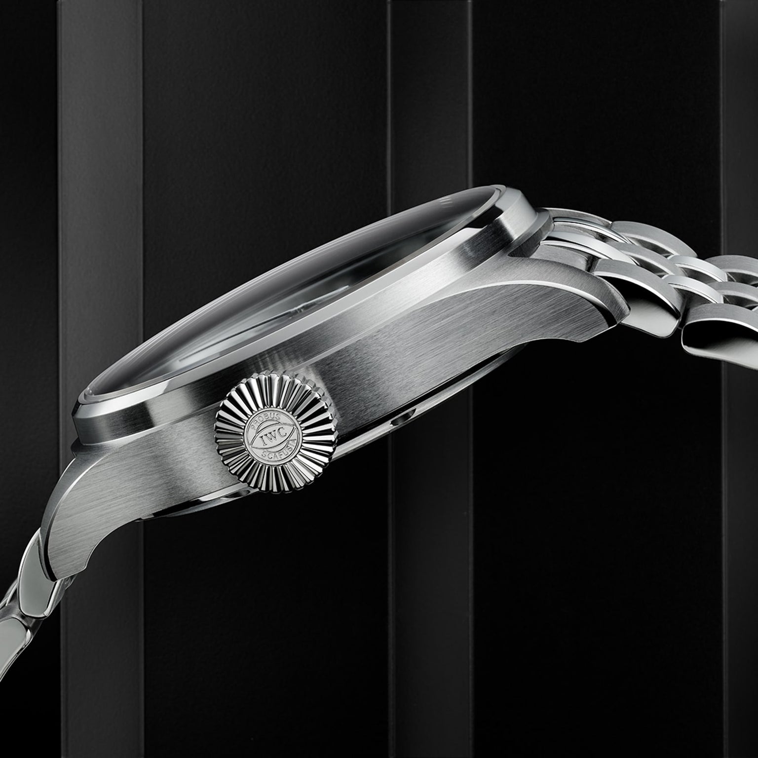 A close-up side view of the IWC Schaffhausen Big Pilots Watch 43, highlighting its textured crown with IWC logo and classic stainless steel case, set against a dark vertically striped background.