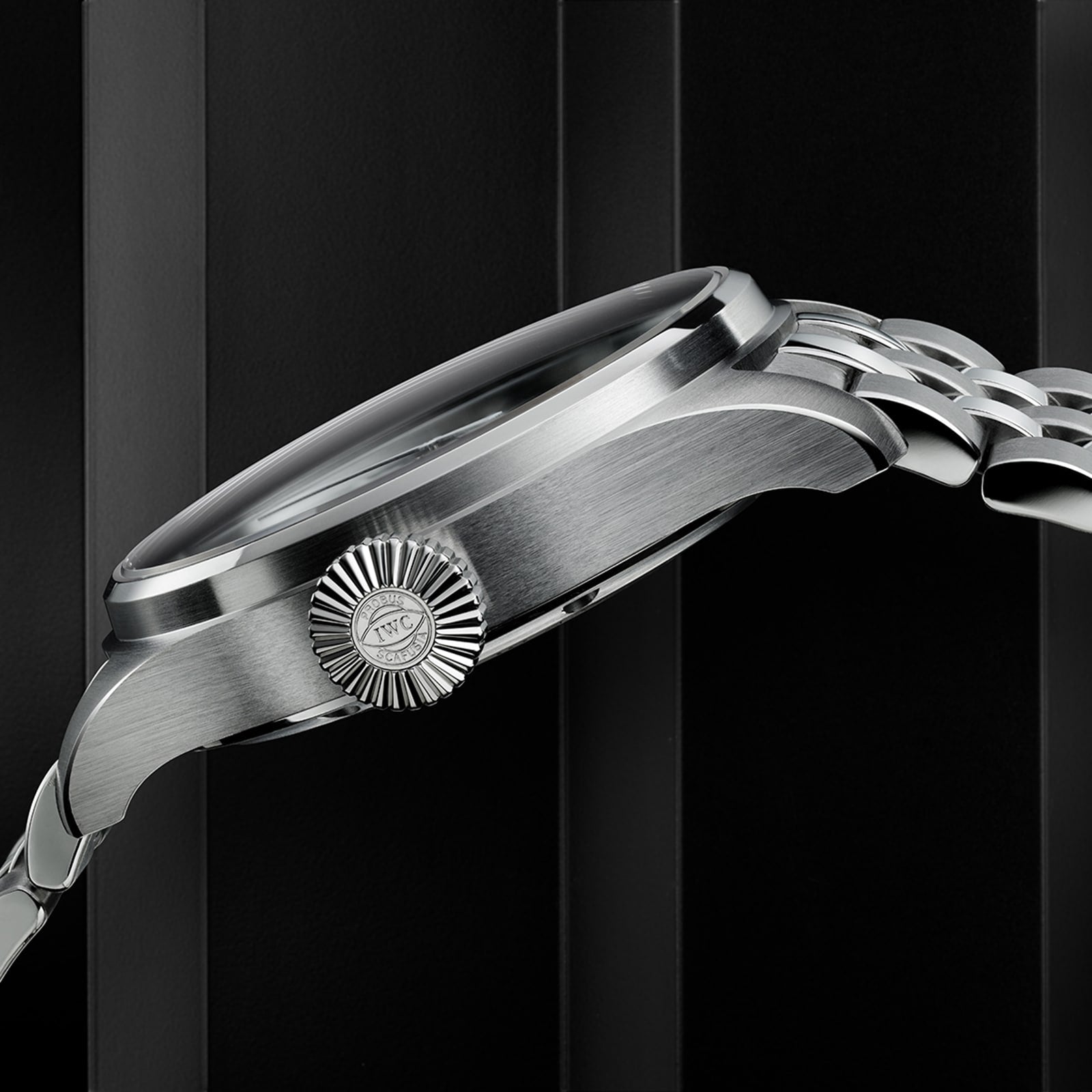 A close-up side view of the IWC Schaffhausen Big Pilots Watch 43, highlighting its textured crown with IWC logo and classic stainless steel case, set against a dark vertically striped background.