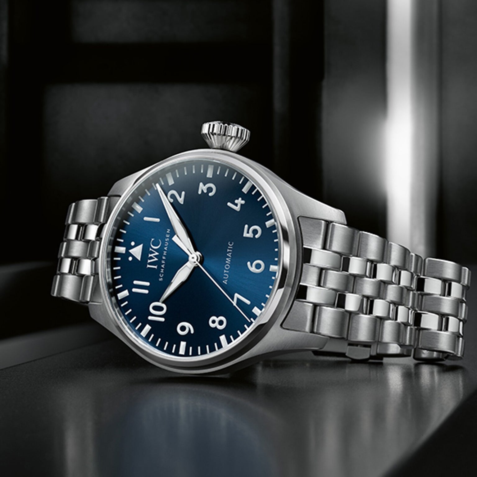 The IWC Schaffhausen Big Pilots Watch 43, featuring a stainless steel case, blue dial with white numerals, and metal link bracelet, is angled on a dark surface, capturing the iconic style of IWC Pilot’s Watches.