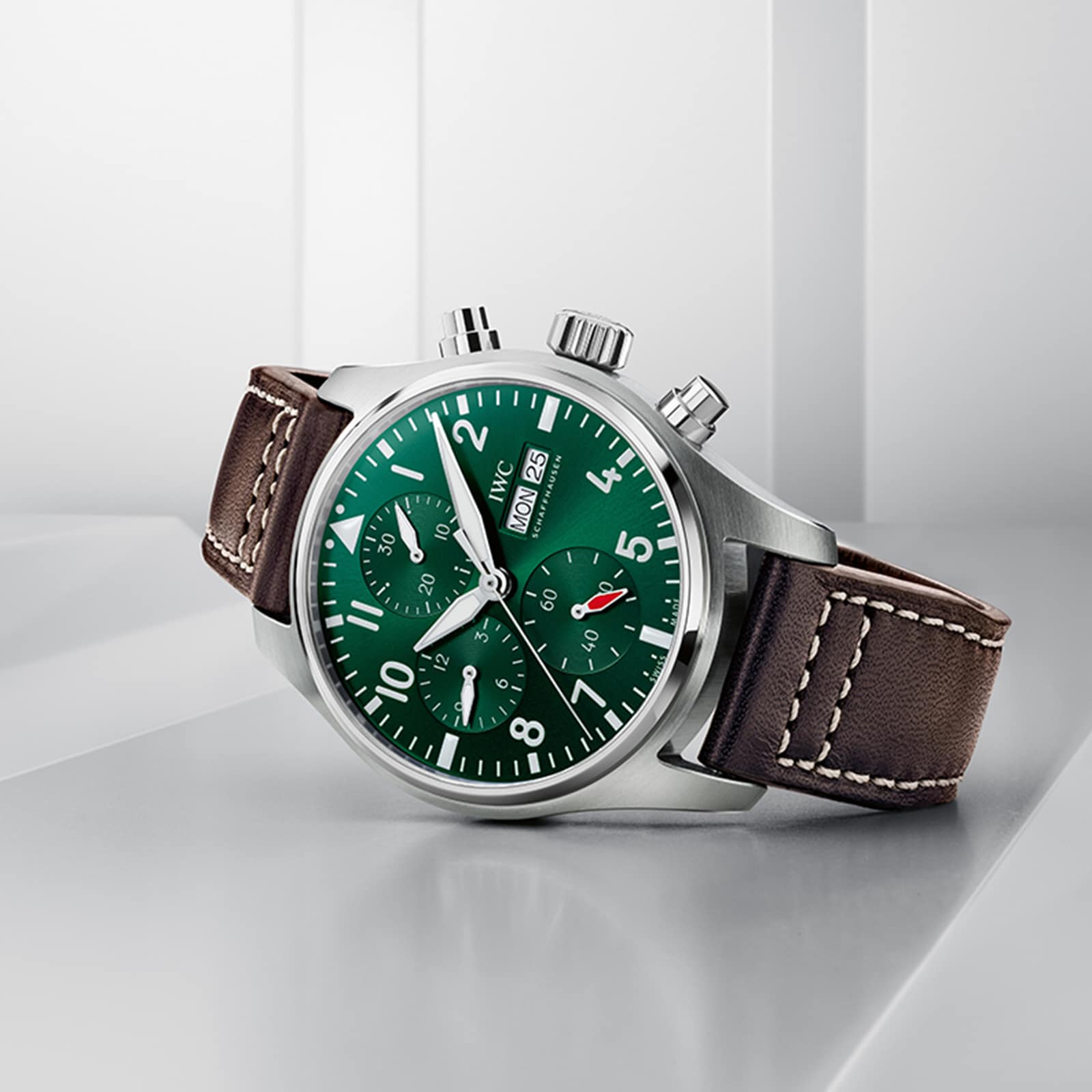 The IWC Schaffhausen Pilots Watch Chronograph 41 features an ergonomic stainless steel case, green dial, brown leather strap, three subdials, and day-date display, elegantly set on a white geometric surface.
