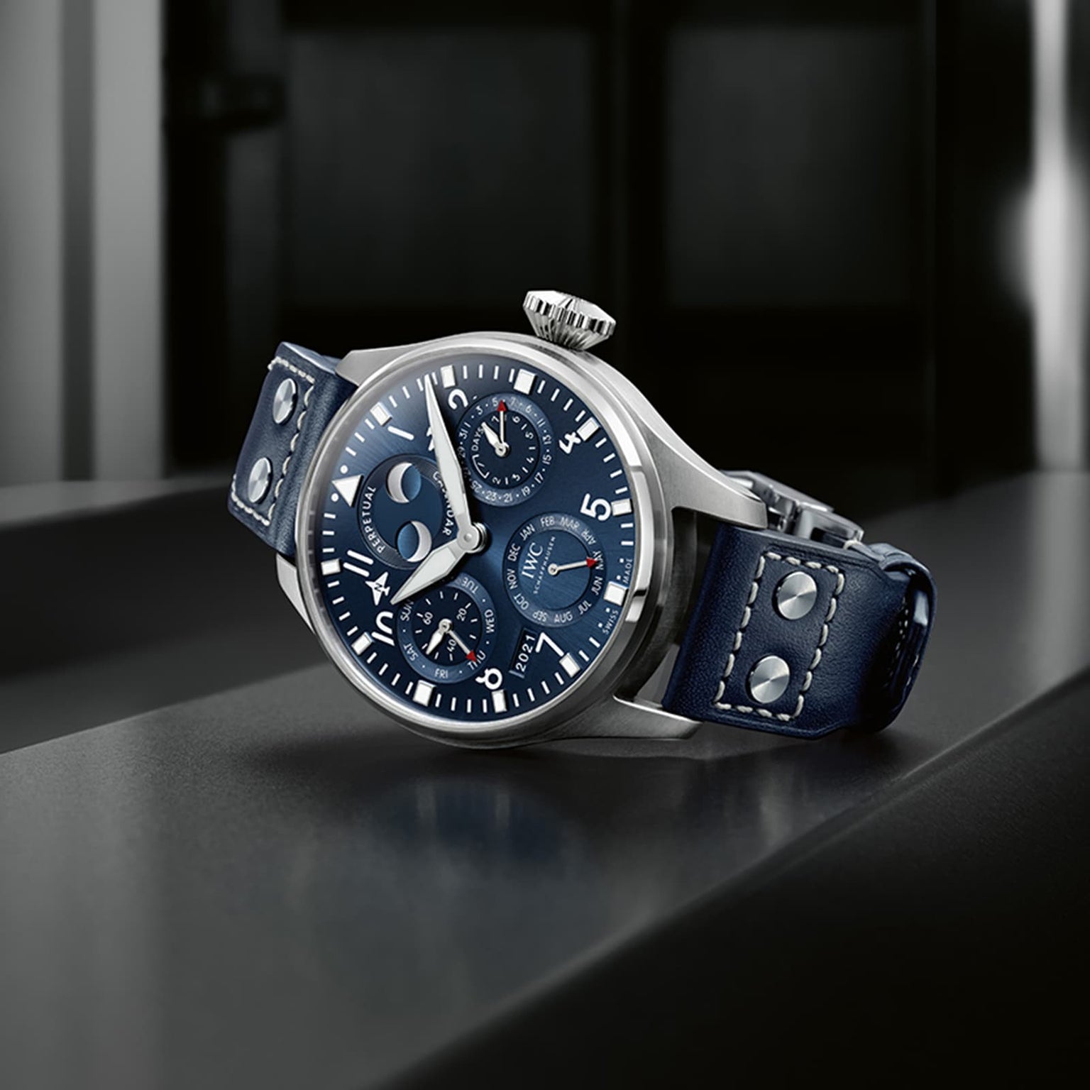The IWC Schaffhausen Big Pilots Watch Perpetual Calendar, with a silver and blue dial, moon phase indicator, and dark blue riveted leather strap, rests on a reflective surface.